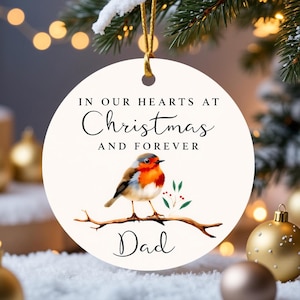 May include: A round Christmas ornament with a robin perched on a branch. The ornament has the text "IN OUR HEARTS AT Christmas AND FOREVER Dad". The bird has a red breast and is set against a white background.