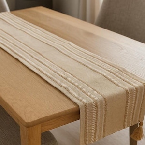 Puede incluir: Un camino de mesa rectangular de color beige con una textura tejida, rayas verticales de color crema y borlas decorativas en cada esquina. La tela presenta un patrón de tejido uniforme y se extiende sobre una mesa de comedor de madera clara.