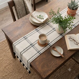 May include: A cream-colored table runner with black plaid stripes and black tassels at the ends. The runner is placed on a wooden table with a ceramic mug on a coaster, a small plate with a spoon, a stack of plates, a potted succulent, and a book.