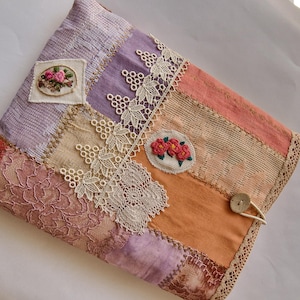Peut inclure: Un objet textile en patchwork aux couleurs orange, violet et rose. Il présente des bordures en dentelle et des motifs floraux brodés. Une fermeture à bouton et boucle est visible. L'objet semble être un étui ou une housse.