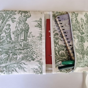 May include: A fabric organiser with a green and white toile pattern, depicting a rural scene. It contains a red book, a spiral notebook, and pens. White ribbon ties secure the organiser.