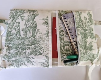 Könnte beinhalten: Ein Stoff-Organizer mit grün-weißem Toile-de-Jouy-Muster, das eine ländliche Szene zeigt. Er enthält ein rotes Buch, ein Spiralnotizbuch und Stifte. Weiße Schleifenbänder sichern den Organizer.