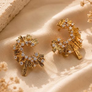 Könnte beinhalten: Ein Paar goldfarbene Ohrringe mit Sonnenstrahl-Design. Jeder Ohrring hat eine gebogene Form mit kleinen, funkelnden Kristallen und radialen Linien. Die Ohrringe werden auf einem cremefarbenen Stoff präsentiert.