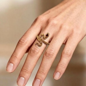 Puede incluir: Un anillo de serpiente dorado con un ojo de gema roja. El anillo está envuelto alrededor de un dedo, mostrando los intrincados detalles del diseño de la serpiente. Las escamas del anillo tienen textura, y la gema añade un toque de color.