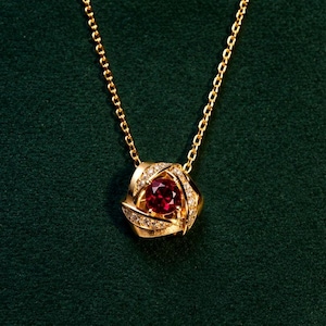 Könnte beinhalten: Eine goldene Halskette mit einem Anhänger, der einen zentralen runden roten Edelstein besitzt, umgeben von einem geschwungenen Metallrahmen mit kleinen klaren Steinen. Der Anhänger hängt an einer zarten Goldkette für ein elegantes Design.