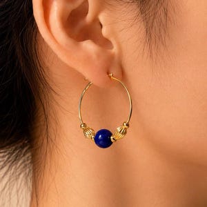 Può includere: Un orecchino a cerchio dorato con una perla sferica centrale liscia di colore blu scuro, affiancata da due perline cilindriche più piccole di colore dorato con superficie scanalata. L'orecchino è formato da un sottile cerchio con chiusura.