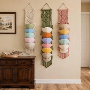 Puede incluir: Tres tapices de macramé verticales con patrones de cuerda trenzada y varillas de madera, diseñados para sostener cinco gorras de béisbol cada uno. Los tapices son de color crema, verde oliva y rosa viejo, con gorras de colores apiladas.