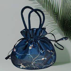 May include: A dark navy blue drawstring handbag featuring a rounded silhouette, two matching top handles, and a delicate embroidered floral vine pattern in shades of orange, light blue, and white across the front surface of the fabric.