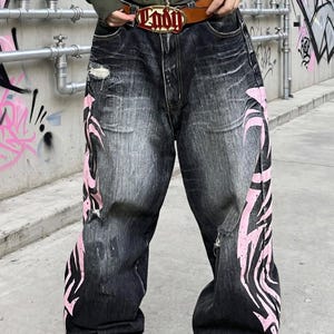 Puede incluir: Vaqueros negros holgados de mezclilla con acabado desgastado, roturas deshilachadas y grandes estampados gráficos tribales de color rosa en los laterales de las piernas. Se sujetan con un cinturón de cuero marrón con una hebilla roja y dorada.