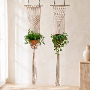 May include: Two off-white macrame wall hangings suspended from wooden dowels. The left piece features a woven heart pattern, while the right has a triangular design. Each holds a terracotta pot containing a green plant with long tassels below.