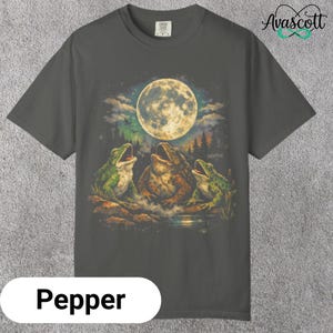 Peut inclure: T-shirt de couleur poivre avec une illustration fantaisiste de trois grenouilles chantant sous la pleine lune. Le motif comprend un ciel nocturne détaillé et une scène de forêt. Le mot "Avascott" est en haut à droite.