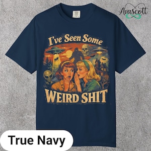 May include: True navy blue t-shirt with a retro graphic design. The design features two women whispering, with UFOs, a skull, and a Bigfoot-like creature in the background. Text reads "I've Seen Some Weird Shit."