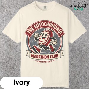 May include: Ivory-colored t-shirt featuring a cartoon illustration of a running mitochondria. The design includes the text "THE MITOCHONDRIA MARATHON CLUB" and "FUELED BY ATP". The graphic is in red, white, and black.