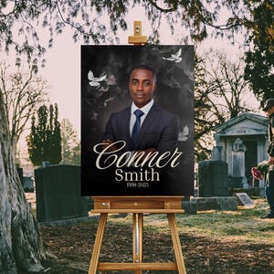 May include: A memorial portrait of Conner Smith, displayed on a wooden easel in a cemetery. The portrait features a man in a suit, with the name "Conner Smith" and dates "1998-2025" below. White doves are depicted in the background.