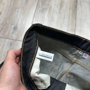 May include: Close-up of dark wash denim jeans. The jeans have a white tag with the text "badfriend" and a small American flag tag. The stitching is a contrasting gold color.