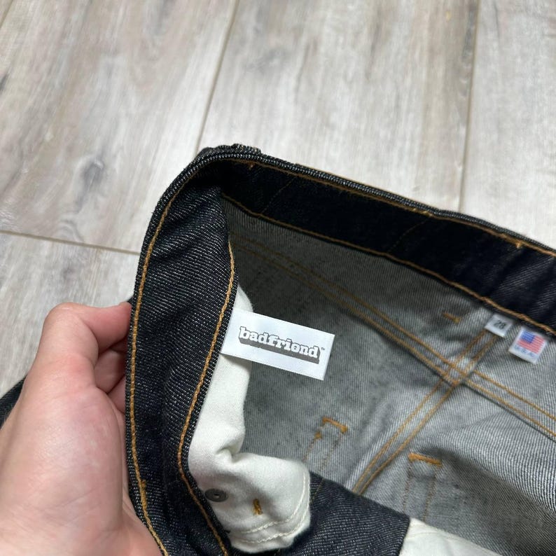 May include: Close-up of dark wash denim jeans. The jeans have a white tag with the text "badfriend" and a small American flag tag. The stitching is a contrasting gold color.