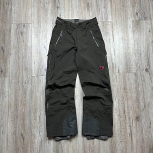 May include: Dark olive-green snow pants with multiple zippered pockets and reinforced patches at the knees and ankles. A red logo is visible on the right leg. The pants have a built-in belt and are designed for winter sports.