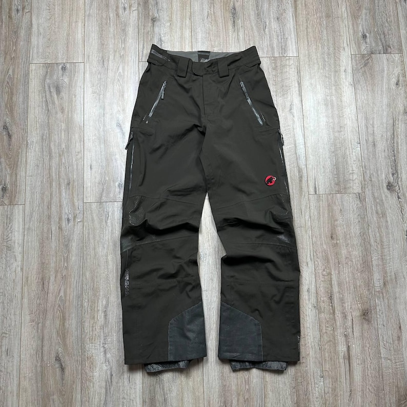 May include: Dark olive-green snow pants with multiple zippered pockets and reinforced patches at the knees and ankles. A red logo is visible on the right leg. The pants have a built-in belt and are designed for winter sports.