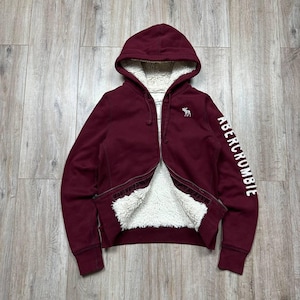 May include: A burgundy zip-up hoodie with a white sherpa lining. The hoodie features a hood, a small embroidered moose logo on the chest, and the word "Abercrombie" printed in white on the sleeve.