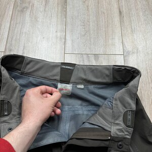 May include: Close-up of dark gray outdoor pants with the brand name "Mammut" visible. The pants have a gray interior lining and a tag with size information. The pants are made of a durable, water-resistant material.