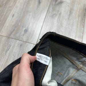 May include: Close-up of dark denim jeans with a white tag. The tag has text that is partially visible. The jeans have yellow stitching and a classic five-pocket design. The denim appears to be a dark wash.