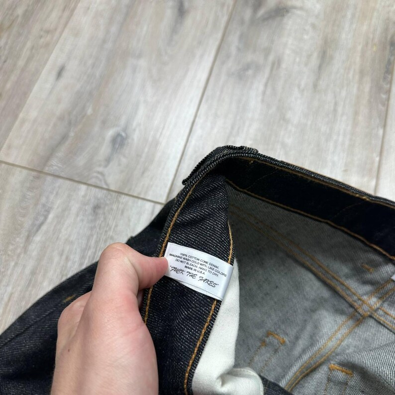 May include: Close-up of dark denim jeans with a white tag. The tag has text that is partially visible. The jeans have yellow stitching and a classic five-pocket design. The denim appears to be a dark wash.