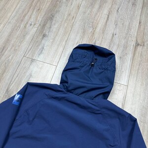 Może przedstawiać: Granatowa kurtka z kapturem ze sznurkiem i małą naszywką z logo na rękawie. Kurtka wykonana jest z lekkiego materiału. Kaptur jest podniesiony, a kurtka jest częściowo rozpięta. Prosty design.