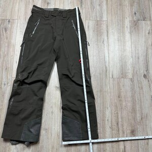 May include: Dark olive-green ski pants with multiple zippered pockets and reinforced patches on the knees and lower legs. A measuring tape is placed next to the pants. The pants have a red logo on the right leg.