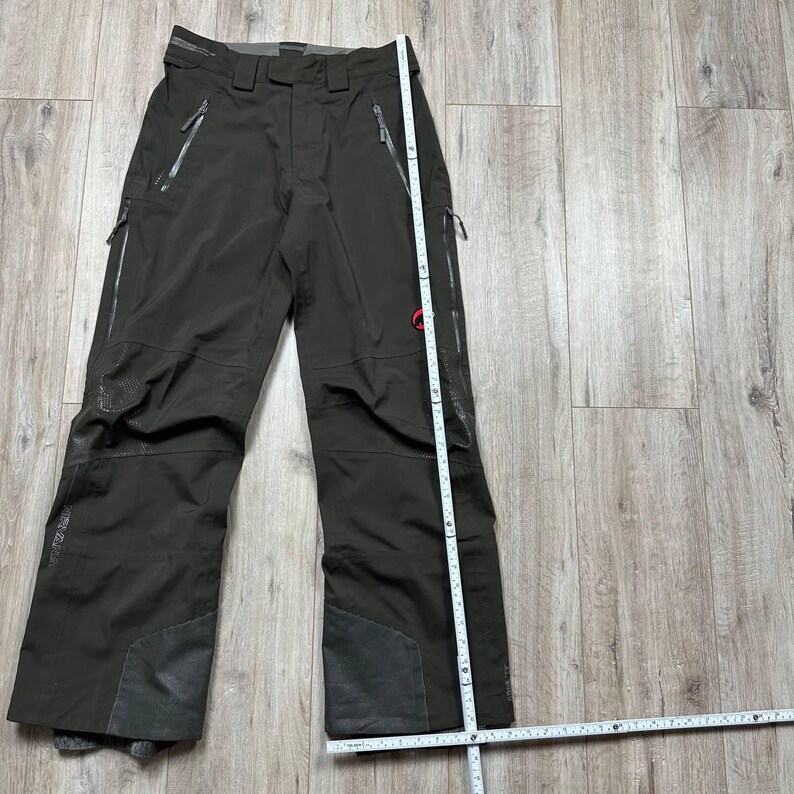 May include: Dark olive-green ski pants with multiple zippered pockets and reinforced patches on the knees and lower legs. A measuring tape is placed next to the pants. The pants have a red logo on the right leg.