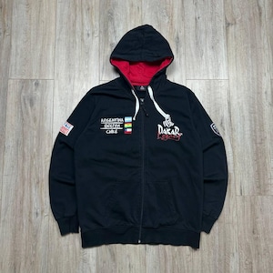 Könnte beinhalten: Ein schwarzer Kapuzenpullover mit Reißverschluss, rotem Innenfutter und weißen Kordeln. Auf der Vorderseite steht "ARGENTINA BOLIVIA CHILE" mit den entsprechenden Flaggen und einem "DAKAR LEGACY"-Grafikdruck. Ein "HERITAGE 1979"-Aufnäher befindet sich am rechten Ärmel.