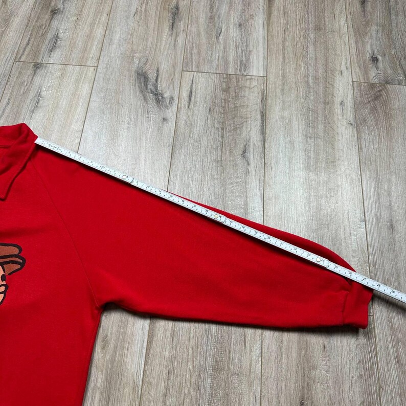 Pu&ograve; includere: Una felpa rossa brillante a maniche lunghe con una grafica a cartoni animati sul davanti. Un metro a nastro &egrave; appoggiato sulla manica, che mostra la lunghezza. La felpa ha un collo.