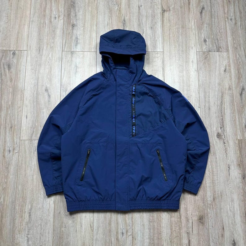 Może przedstawiać: Granatowa kurtka z kapturem, z pełnym zapięciem na zamek i dwiema przednimi kieszeniami. Kurtka ma sznurek w talii i małe logo na lewym rękawie. Wykonana z lekkiego materiału.