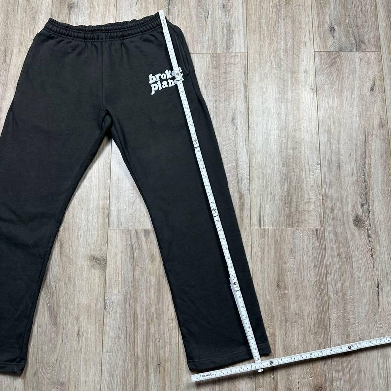 Może przedstawiać: Czarne spodnie dresowe z białym napisem "broken planet" na lewej g&oacute;rnej nogawce. Spodnie mają elastyczną talię i prostą nogawkę. Wzdłuż boku spodni umieszczono miarkę.