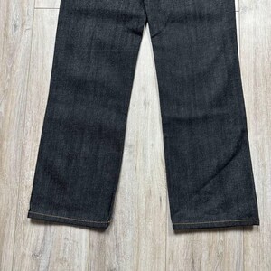 May include: Dark wash denim jeans with a straight leg cut. The jeans have a classic five-pocket design and a button and zipper closure. The hem is finished with a contrasting yellow stitch.