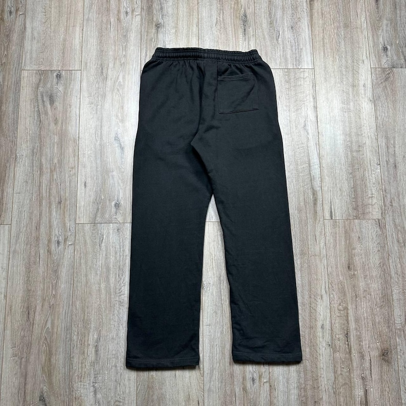 Może przedstawiać: Czarne spodnie dresowe z elastyczną talią i jedną tylną kieszenią. Spodnie wykonane są z miękkiego materiału i mają luźny kr&oacute;j. Zdjęcie przedstawia tył spodni, ułożonych płasko na drewnianej powierzchni.