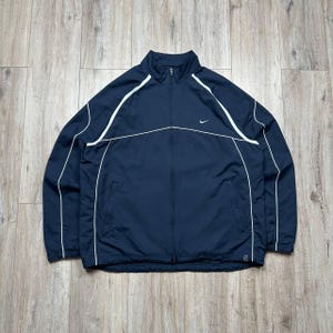 May include: A navy blue zip-up track jacket featuring a high collar, long sleeves, and white piping accents along the shoulders and chest. A small white Nike swoosh logo is embroidered on the upper left chest, and the jacket includes two side pockets.