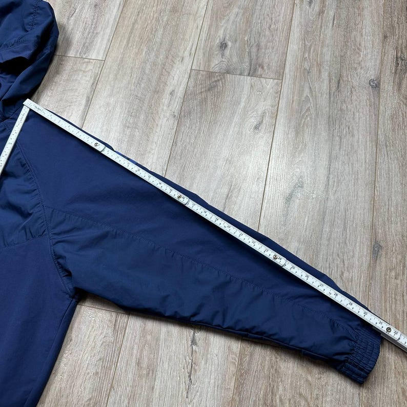 Może przedstawiać: Granatowa kurtka z kapturem i zapięciem na zamek błyskawiczny. Taśma miernicza jest ułożona na rękawie, pokazując długość około 76 cm. Kurtka ma prążkowany mankiet i kaptur ze sznurkiem.