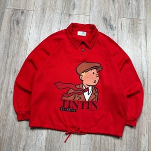 Pu&ograve; includere: Una felpa rossa a maniche lunghe con una grafica a cartoni animati di Tintin, con la scritta "TINTIN REPORTER" stampata sotto. La felpa ha un collo e una vita regolabile con coulisse.