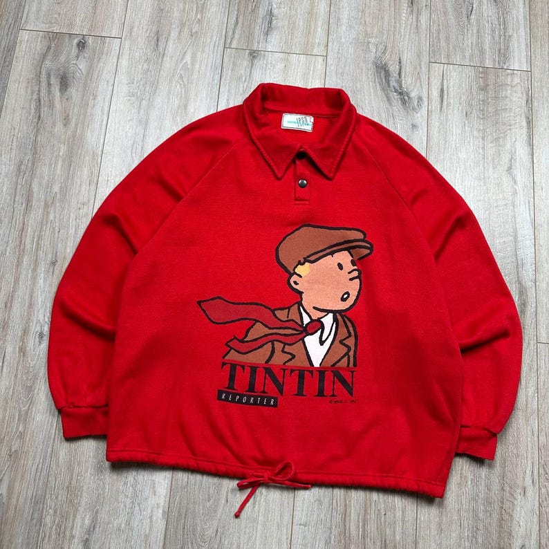 Pu&ograve; includere: Una felpa rossa a maniche lunghe con una grafica a cartoni animati di Tintin, con la scritta "TINTIN REPORTER" stampata sotto. La felpa ha un collo e una vita regolabile con coulisse.