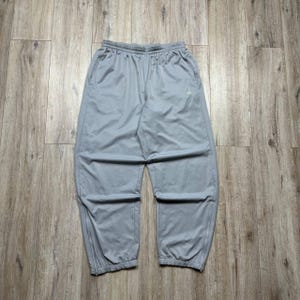 May include: Light grey athletic trousers featuring an elasticated waistband, side pockets, and a small logo on the upper right thigh. The trousers have a relaxed fit with tapered legs that end in zippered cuffs, providing a functional and clean appearance.