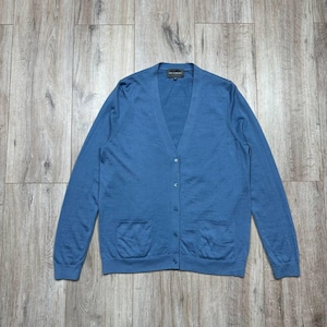 May include: A blue cardigan with a V-neck, long sleeves, and two front pockets. The button-down sweater is made of a soft material and is displayed on a wooden surface. The cardigan is a solid color.