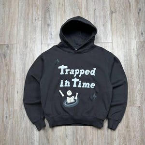Może przedstawiać: Czarna bluza z kapturem z białym napisem "Trapped in Time" oraz grafiką szkieletu w okręgu. Bluza posiada r&oacute;wnież zdobienia w kształcie gwiazd.