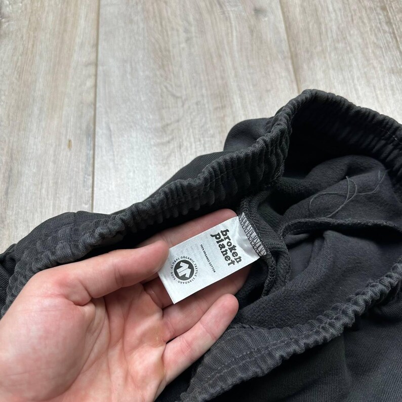 Może przedstawiać: Zbliżenie ciemnoszarych spodni dresowych z elastycznym pasem. Widoczna jest biała metka z napisem "Broken Planet". Etykieta zawiera r&oacute;wnież logo i słowa "100% Organic Cotton".