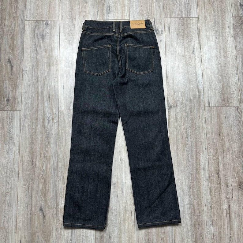 May include: Dark wash denim jeans with a straight leg cut. The jeans have two back pockets and a small leather patch on the back waistband. The stitching is a contrasting gold color.