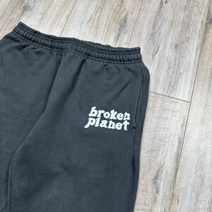 Może przedstawiać: Ciemnoszare spodnie dresowe z elastyczną talią i boczną kieszenią. Słowa "broken planet" są wydrukowane na lewej nogawce białym, stylizowanym krojem pisma. Spodnie są wykonane z miękkiego materiału.