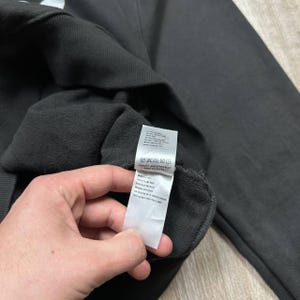 Może przedstawiać: Ciemnoszara bluza z dołączoną białą metką. Etykieta zawiera instrukcje pielęgnacji i tekst. Bluza wykonana jest z miękkiego materiału. Zdjęcie zrobione jest z bliska.