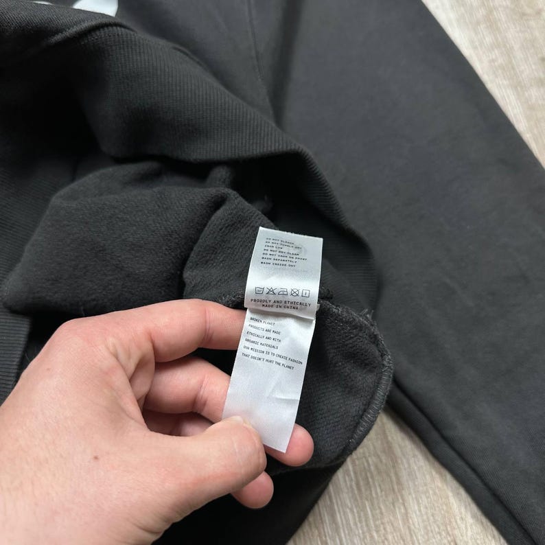 Może przedstawiać: Ciemnoszara bluza z dołączoną białą metką. Etykieta zawiera instrukcje pielęgnacji i tekst. Bluza wykonana jest z miękkiego materiału. Zdjęcie zrobione jest z bliska.