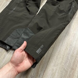 May include: Close-up of dark brown outdoor pants with reinforced patches and the text "GORE-TEX Soft Shell". The pants appear to be made of a durable, water-resistant material, suitable for outdoor activities.