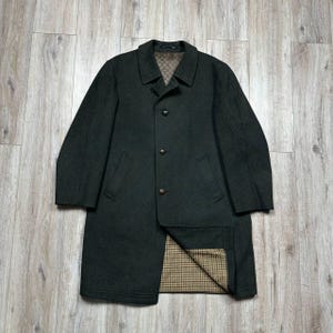 Puede incluir: Un abrigo de lana verde oscuro con un diseño clásico. El abrigo tiene cuello con solapas, tres botones y dos bolsillos delanteros. El forro interior es un estampado a cuadros marrón y beige. El abrigo está sobre una superficie de madera clara.