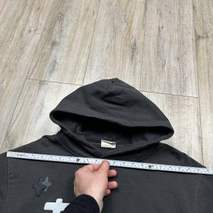 Może przedstawiać: Ciemnoszara bluza z kapturem z kapturem i białą metką. Bluza z kapturem ma biało-szary wz&oacute;r z przodu. Linijka jest trzymana na bluzie z kapturem, aby zmierzyć jej szerokość.